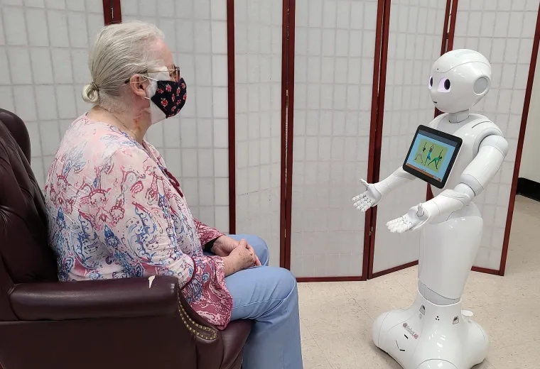 a white android about 4 feet tall interacts with a seated elderly woman