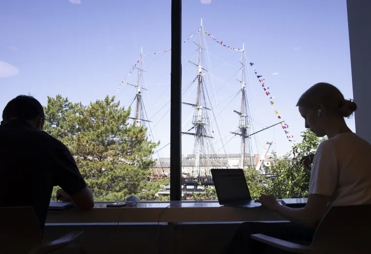 Silhouette of 2 people on laptops in the school in front of a sailing ship outside of the window.
