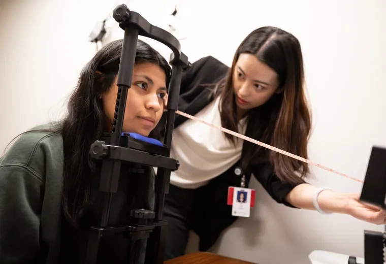 woman rests chin on bar device while another woman measures the distance from her face to a camera