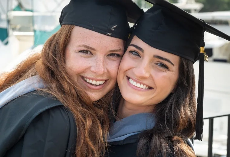 two women beam for the camera wearing caps and gowns in front of boston skyline