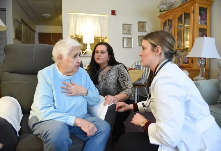 in a living room, an older woman with white hair puts a hand to her chest as two younger females crouch down to speak to her
