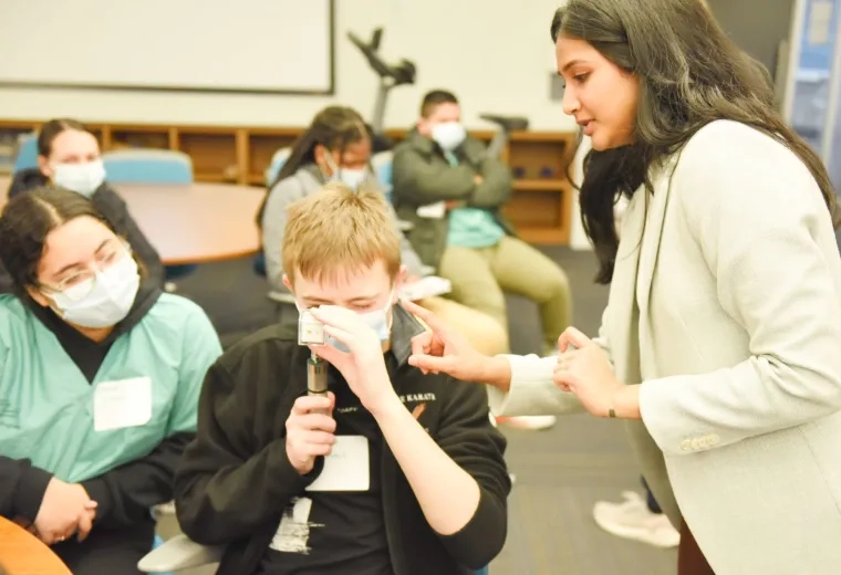 woman in blazer points at a cylindrical device that a man is holding to his eye