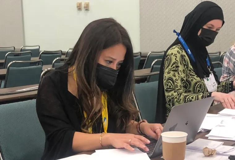 woman sits in chair at laptop next to another woman wearing a hijab