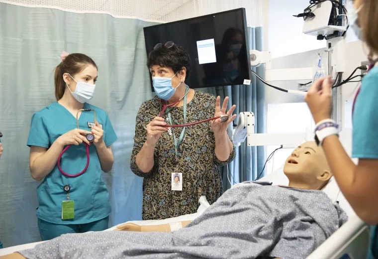 Karen holds a stethoscope and shows a student in scrubs how to apply it to a manikin in a hospital bed