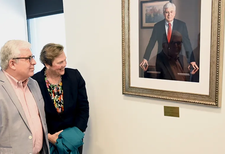 alex in a grey blazer and paula in a black blazer look at a painting of alex in a gold frame on the wall