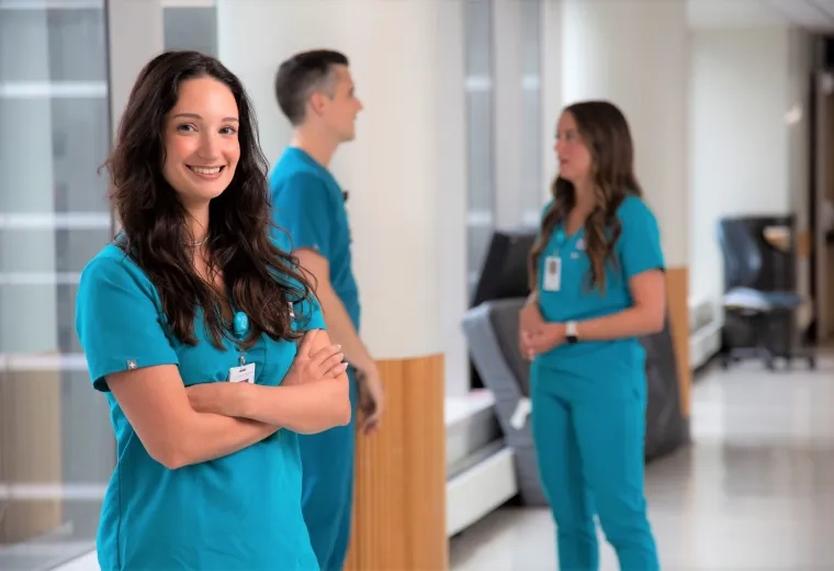 mackenzie has long dark wavy hair and stands with her arms crossed smiling in a hallway