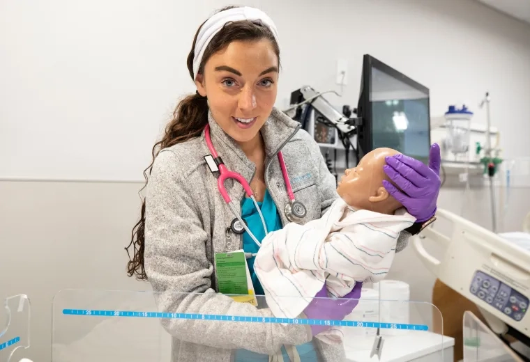 A student in the MGH IHP Fast-Track BSN Program holding an infant mannequin during a hands-on nursing licensure preparation session. The student is dressed in scrubs and is actively participating in the accelerated nursing curriculum, which is part of an intensive nursing training program designed for professionals transitioning to nursing careers.