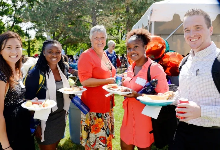 woman in bright orange top with four young-adult looking people in backpacks standing outside on a sunny day with plates of food.