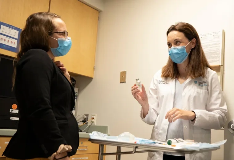person in white lab coat holds up a vial to another person wearing a suit jacket in a doctors office exam room