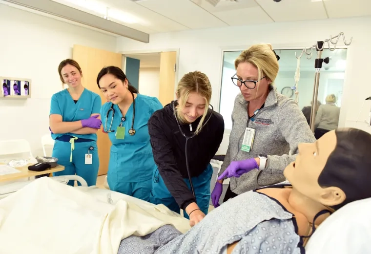 a woman in scrubs takes the pulse of a manikin in a bed while a woman in a MGH fleece looks on