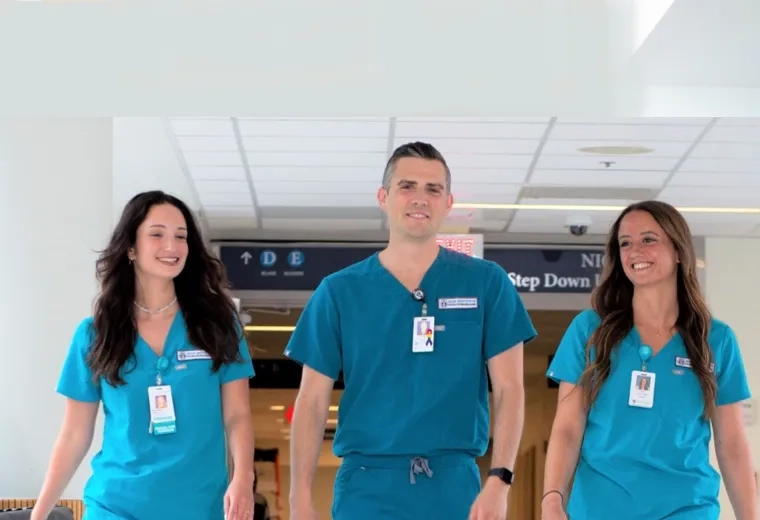 a man and two women in blue scrubs and name badges walk down a hospital hallway (step down unit sign in the background)