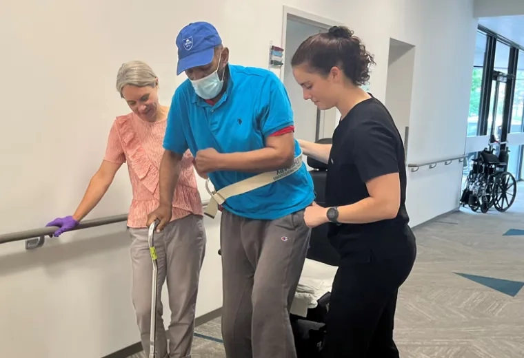man in a surgical mask is helped by two women on either side to walk down a hallway