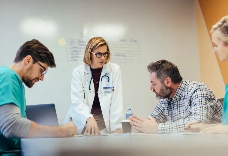 woman in white coat and stethoscope gestures to people at a table wearing scrubs