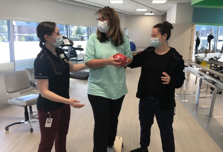 two women assist a third woman balancing on a wooden plank with a red ball in her hand