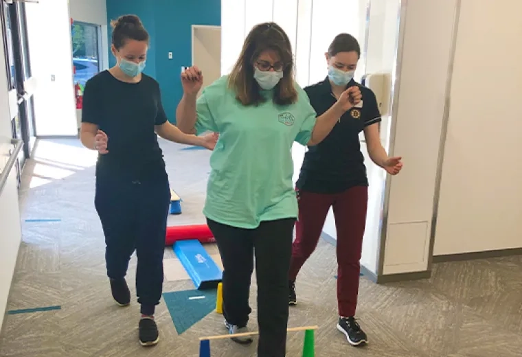 two women assist a third woman stepping over a low stick across two cones