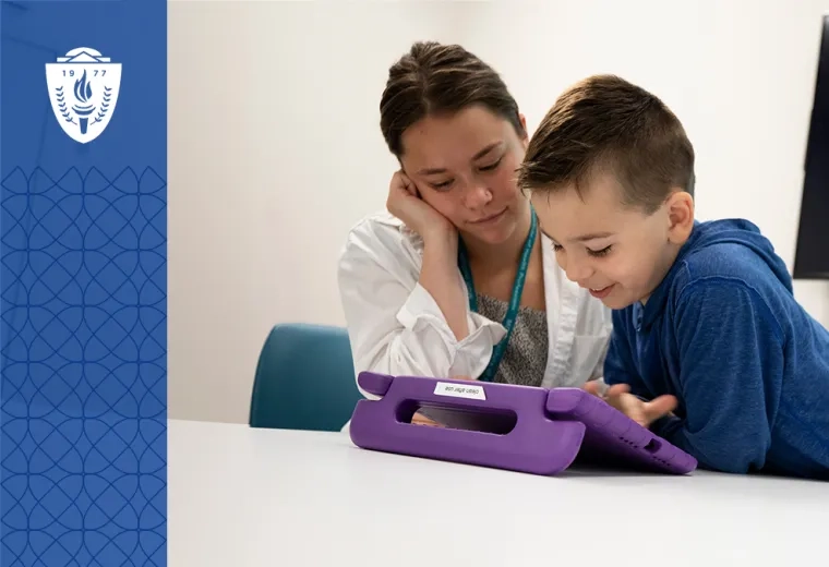 student in a lab coat looks at something on a tablet with a little boy while a colorful cartoon is on the TV in the background