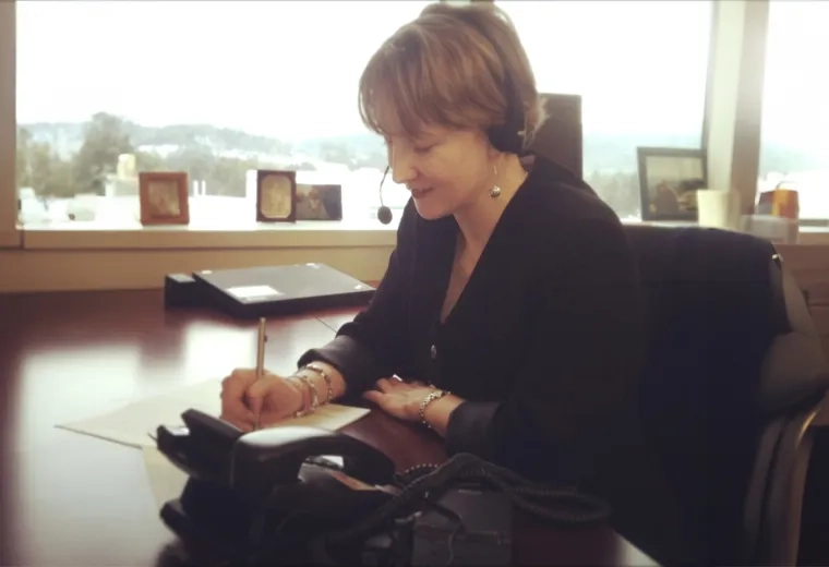 kathy sits at a desk with a headset on and takes notes on a sheet of paper
