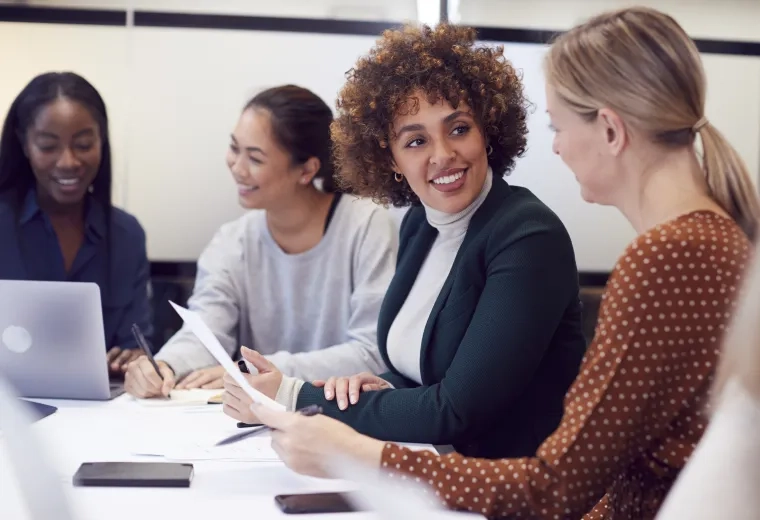 four women in business casual attire sit at a table and discuss paperwork