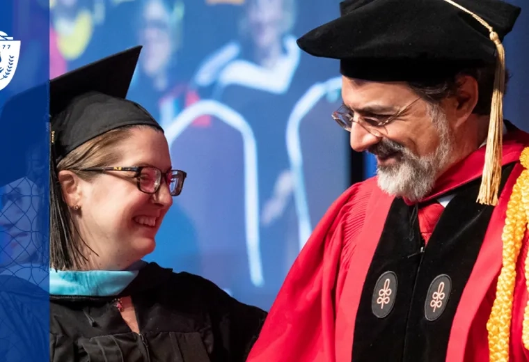 professor in graduaiton regalia (cap and red gown) smiles at a woman graduating who is in blue regalia