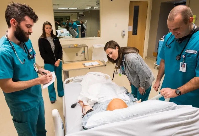 2 men and 2 women in scrubs stand around a manikin in a hospital bed with a big mirrored window in the wall behind them
