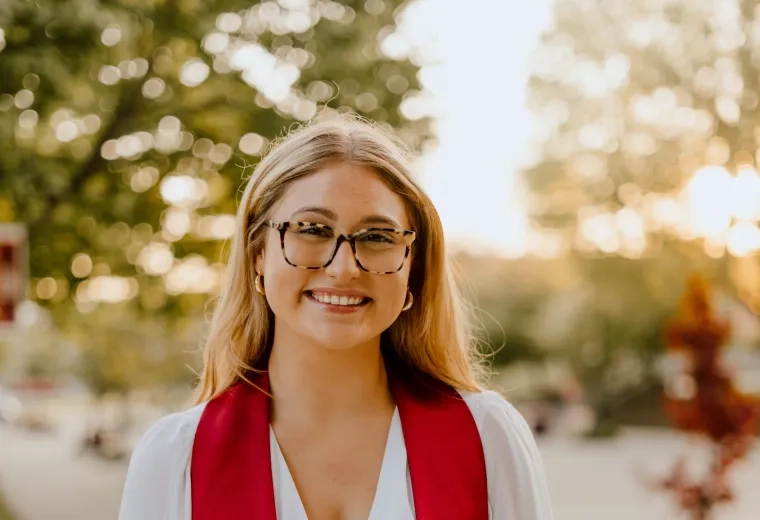 Sydney Zech has blond hair and glasses. She is wearing a white dress with a red graduation ribbon.