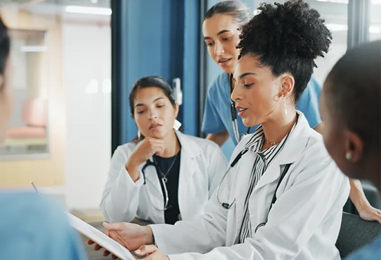 woman in lab coat and stethoscope showing others around the table something on her ipad 