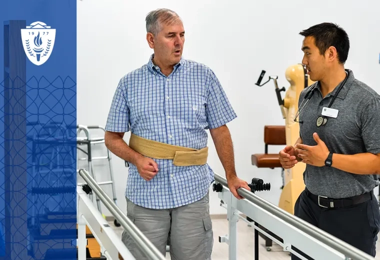 older man with a strap around his waist stands between parallel bars while a young man explains something to him