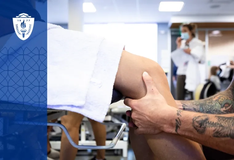 a person sits in front of another who is on a medical table, the focus are the person's hands around the other's knee