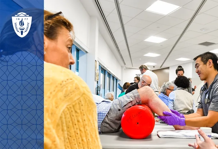 a man helps an older man roll his legs on a red foam tube on a treatment table while two women talk in the foreground