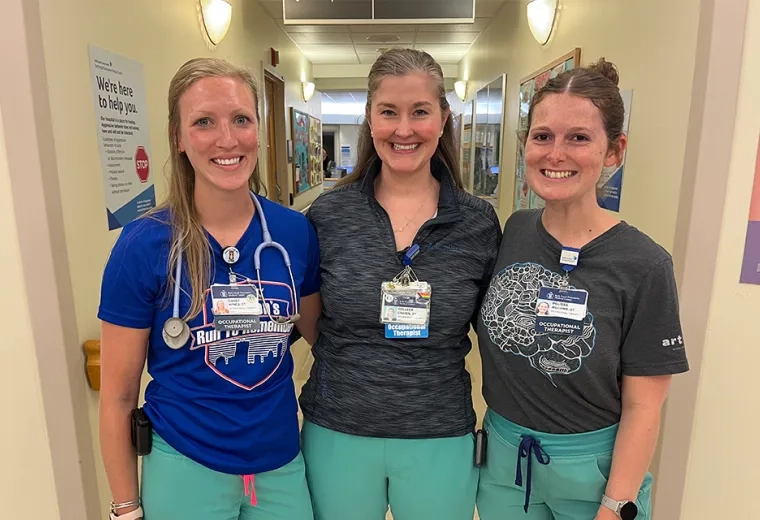 three women in matching scrub bottoms stand in a clinical hallway arm and arm 