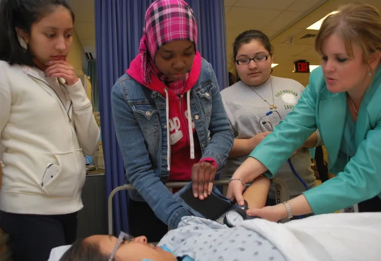 A woman in a hospital setting demonstrates taking a patient's blood pressure to three girls