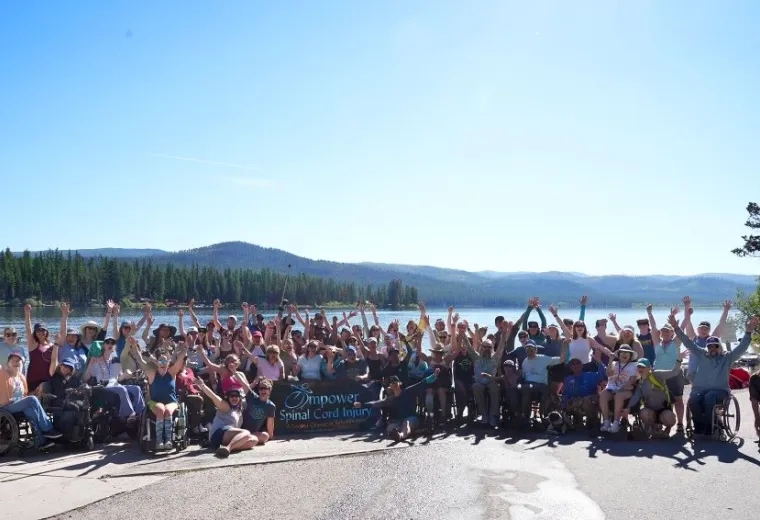 A large group of people stand together for a photograph in front of a lake with trees and mountains in the background