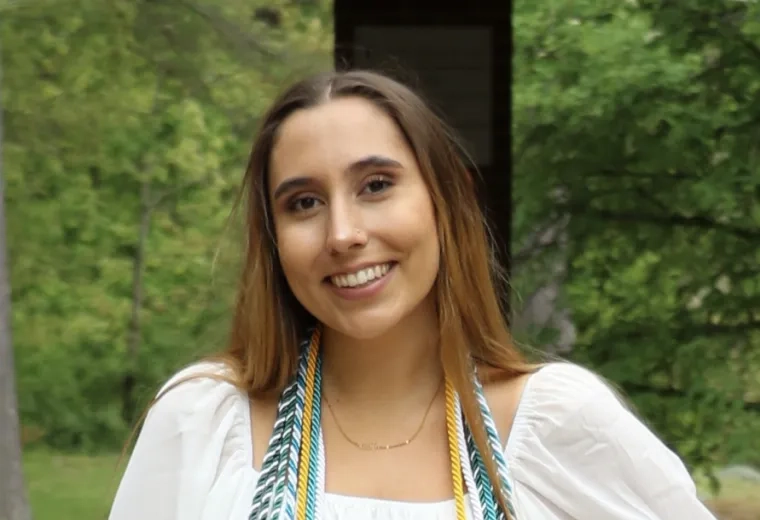 Maegan Mannes is in a white dress with graduation cords and has long brown hair