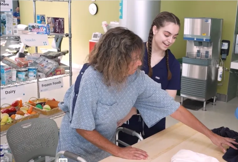 A student wearing hospital scrubs helps a woman wearing a hospital gown practice folding the laundry while a man with an id bade watches