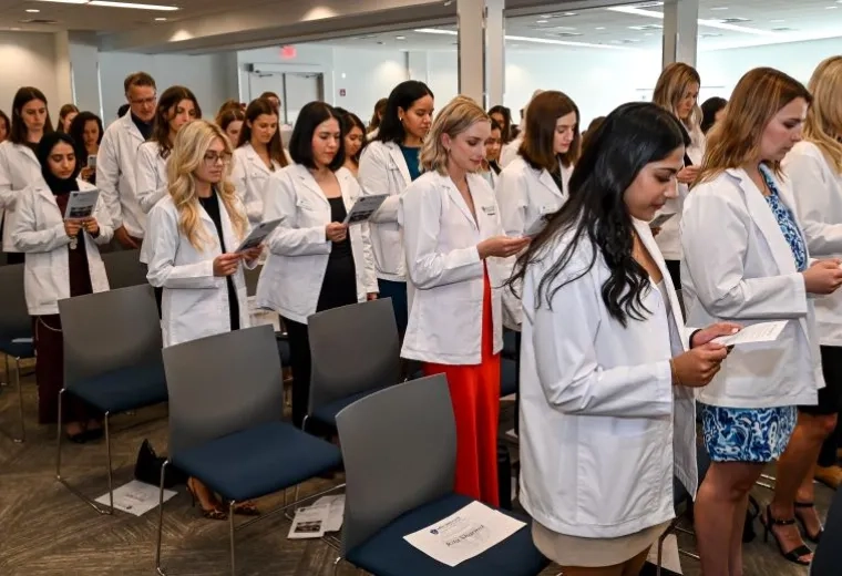 People in an audience, all wearing white coats, read from a piece of paper