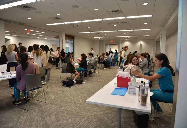 People sit at multiple tables with their arms ready to get a flu shot while nursing students administer them and a crowd waits on the side of the room 