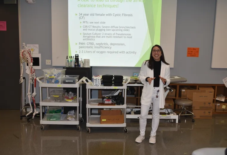 A woman in a white sweater and pants stands in front of a screen in a classroom with medical items behind her