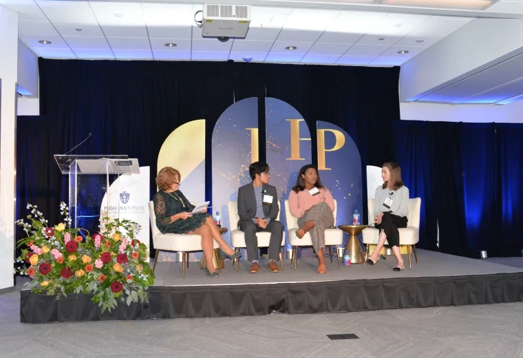 Three women sit in chairs on a stage with a backdrop displaying IHP