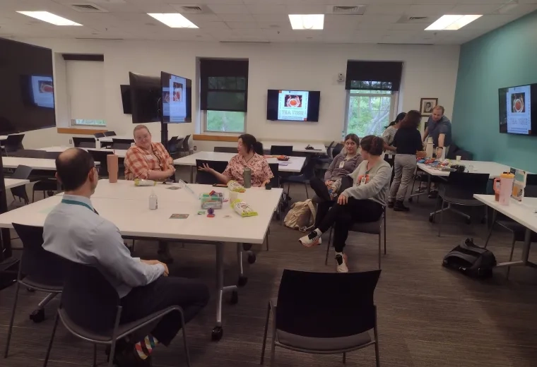 Five people sit around a table while several people in the background stand in front of a table being served food and drinks