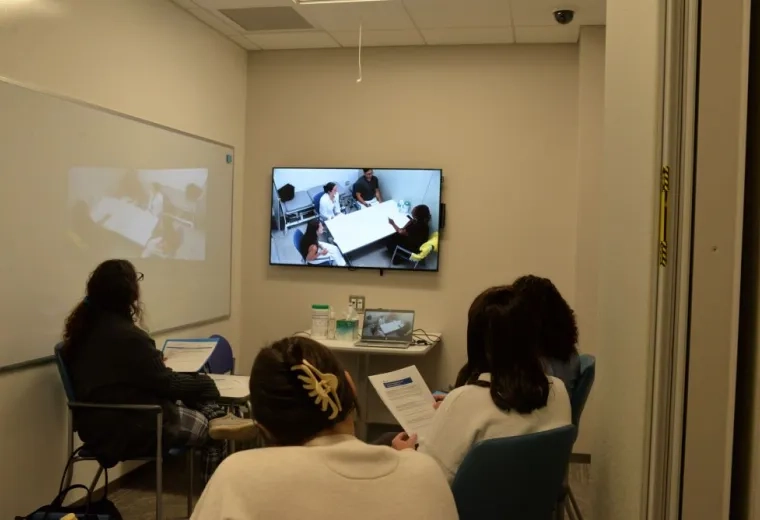 Four people sit in a room watching a screen that shows people sitting around a table