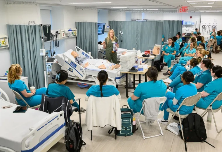 a woman stands in front of a manikin on a hospital bed while people in scrubs sit in a semi circle around her