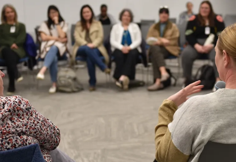A woman holding a microphone is shown from behind while a group of people sit across from her listening