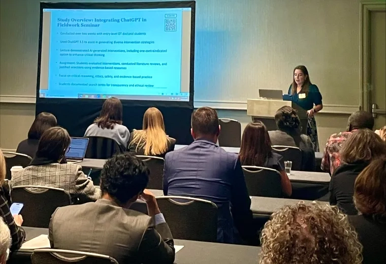 A woman stands at a podium with a screen to her right and an audience sitting in front of her