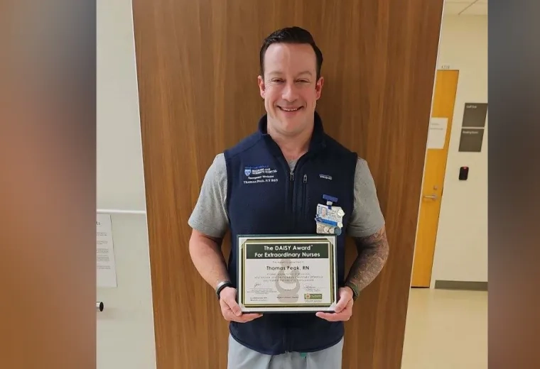 A man stands with his back to a wall holding a certificate for the Daisy Award for Extraordinary Nurses