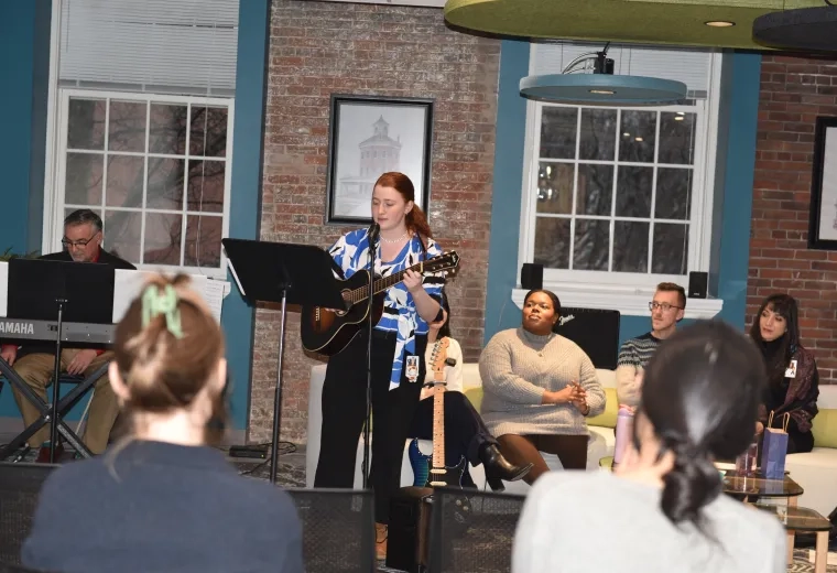 A woman plays the guitar while a man plays the keyboard behind her and four people sit on coach watching from behind her