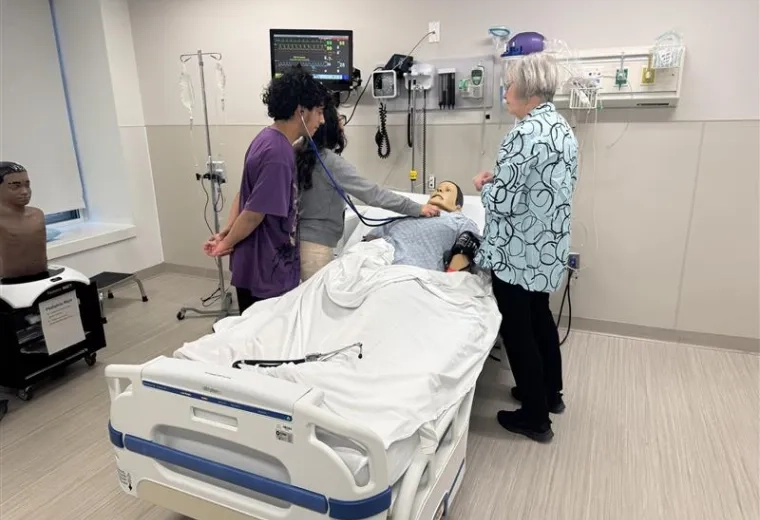 A person holds a stethoscope to a mannikin while another listens to the other end of the stethoscope while a third person looks on. 