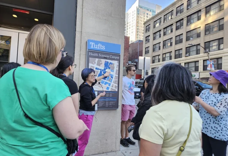 A group of people listen to a woman pointing to a map on a building under a sign that says Tufts