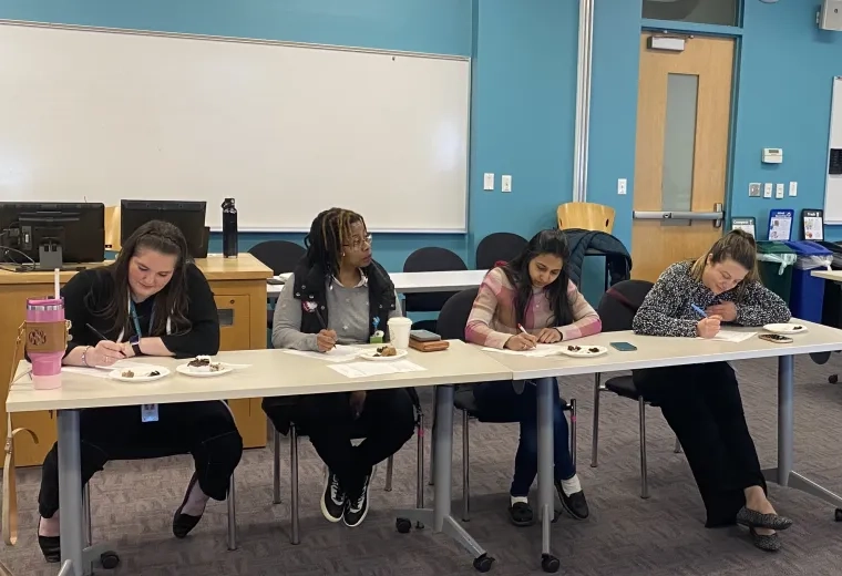 Four women write on paper in front of them while a fifth looks on