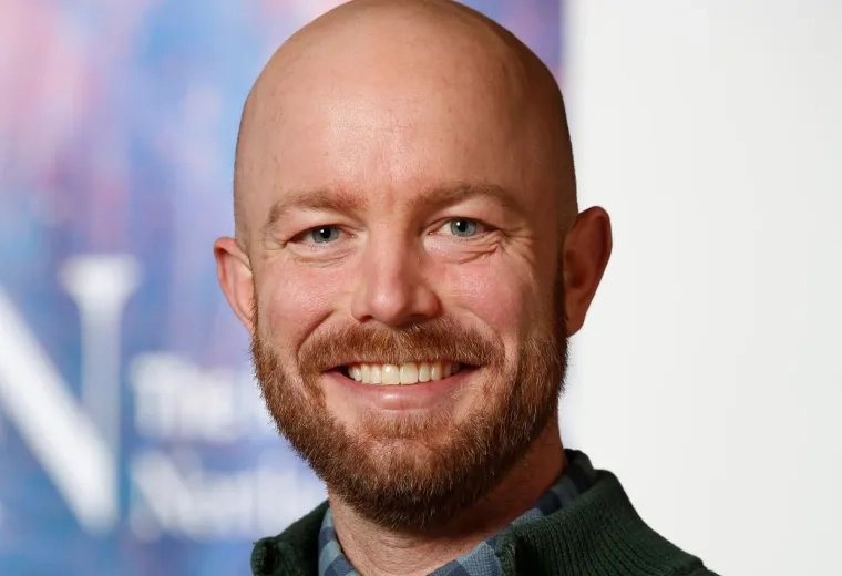 Robert Cavanaugh has a bald head and a brown beard. He is wearing a black sweater over a blue checkered dress shirt.