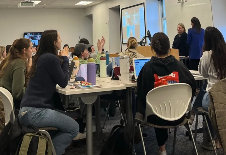 People sit at a table with computers in front of them with two people in the front of the room at a podium